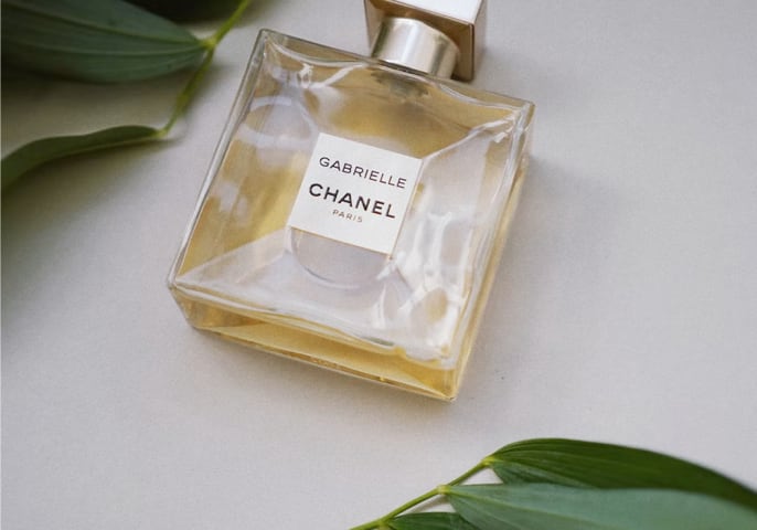 Gabrielle Essense perfume bottel laying flat on a table with green leaves above and below it.